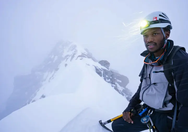 Alpinist with headlamp lit, using ice axe and compass to navigate a snowy alpine ridge in dense fog or blowing snow, facing adverse conditions and alpine-specific challenges.