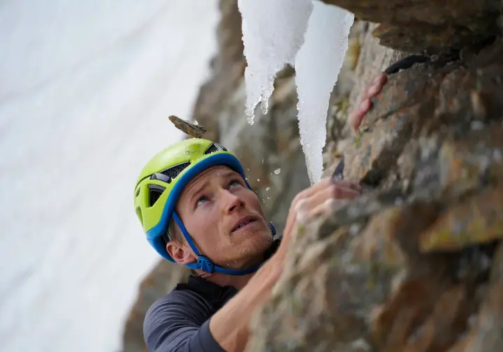 Climber on an alpine route with a small piece of ice deflecting off their helmet, highlighting protection against rockfall and icefall hazards from above.