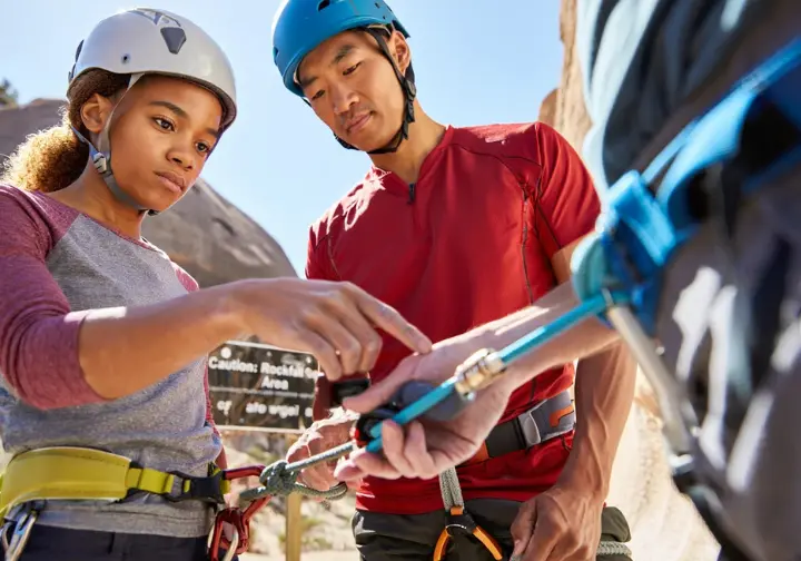 Two diverse climbers meticulously performing a pre-climb partner check (harness, knot, belay device) at an outdoor crag, emphasizing safety protocols and hazard management in sport climbing.