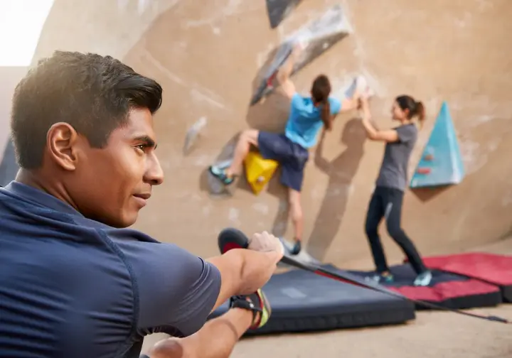 Boulderer performing pre-climb warm-up stretches for injury prevention, with another climber being safely spotted on a well-padded problem in the background, promoting climbing smarter and longer.