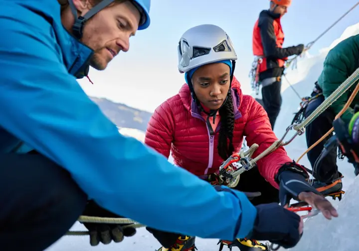 Ice climbing instructor conducting a pre-climb safety check (helmet, harness) with a beginner student at the base of an ice flow, emphasizing critical safety for beginners.