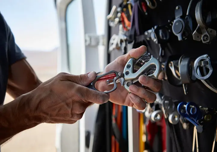 A trad climber's hands perform maintenance on a cam, checking the trigger action and lobes to ensure the protection remains reliable.