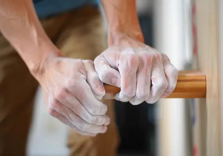Extreme close-up of a boulderer's hands performing a maximal hang on a small crimp hold on a wooden hangboard, illustrating training for forging iron finger strength and tendons.