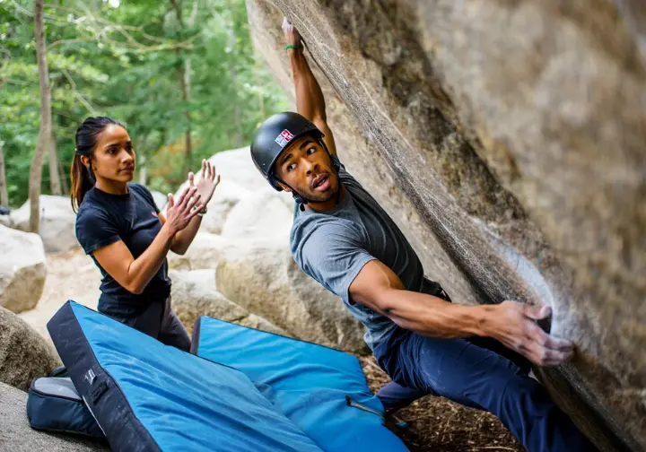 Attentive spotter protecting a climber on an outdoor boulder, with multiple crash pads ensuring a safe landing, illustrating bouldering safety for minimizing risks and maximizing fun.