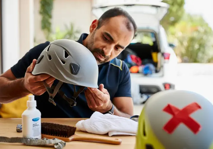 Climber inspecting a climbing helmet for damage, with cleaning supplies nearby and a retired helmet in the background, illustrating helmet lifespan, care, and maintenance.