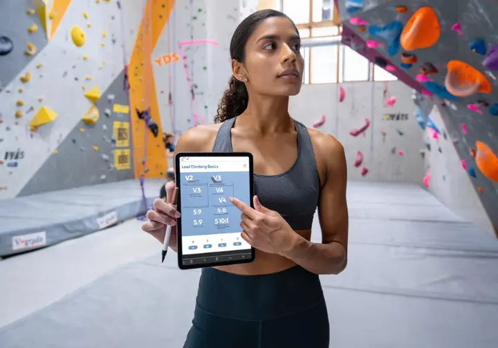 Climber in an indoor gym reviewing a structured training plan on a tablet that includes V-Scale and YDS grade progressions and an introduction to lead climbing, with various gym walls in the background.