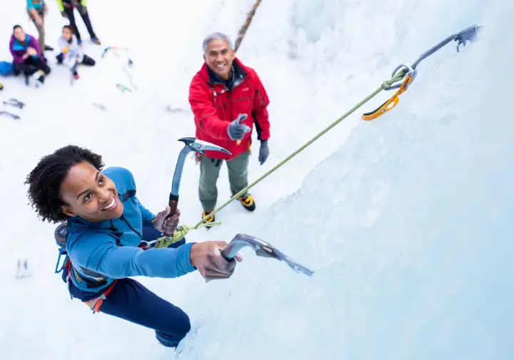 Beginner ice climber smiling after a good tool placement on an ice park wall during an introductory course, with a guide offering encouragement, showing how to get started.