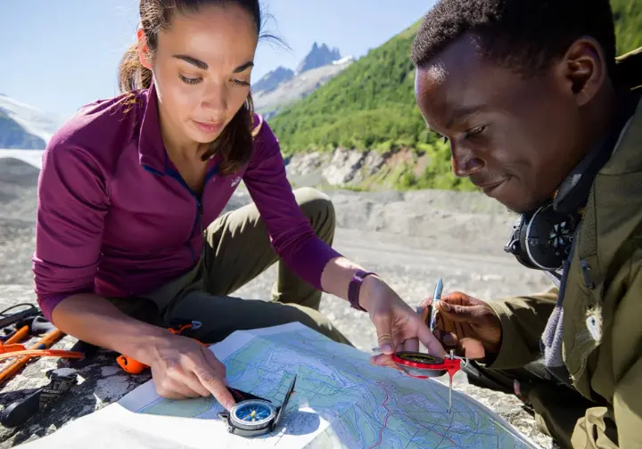 Experienced alpine mentor teaching a newer alpinist map and compass skills in an outdoor training area, cultivating an alpine navigation mindset through continuous learning.