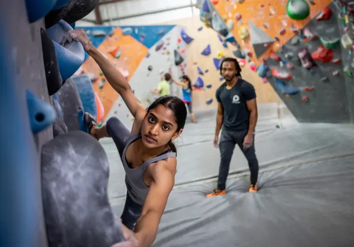 Beginner boulderer making her first moves on an easy indoor climbing wall with large holds and thick mats, with an instructor offering encouragement, illustrating getting started in bouldering.