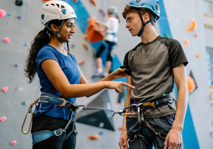 Two diverse climbers diligently performing a partner check, with one guiding the other, to prevent human error from complacency and miscommunication.