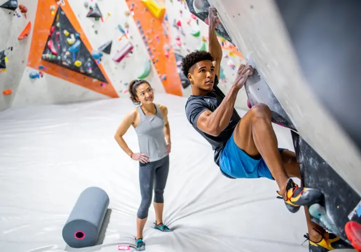 Intermediate climber paused on a challenging indoor boulder problem, reflecting on mistakes, with a coach, foam roller, and better shoes nearby, symbolizing overcoming plateaus and injury prevention.