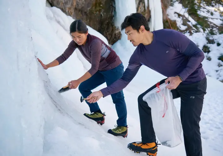 Two ice climbers practicing Leave No Trace at a wilderness ice crag: one gently brushing ice, the other packing out trash, showing respect for the ice and environmental ethics.