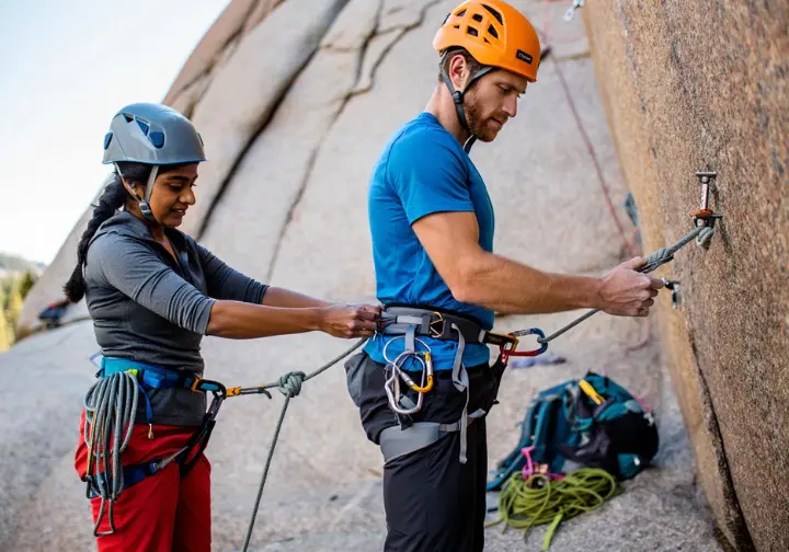 Two diverse trad climbers performing a meticulous pre-climb partner check (harness, knot, belay device) at a crag, emphasizing "Safety First, Always" in trad climbing risk management.