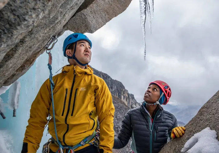 Two diverse mixed climbers at a belay stance in an alpine environment, one inspecting gear placement, the other assessing overhead hazards, demonstrating safety protocols and risk management.