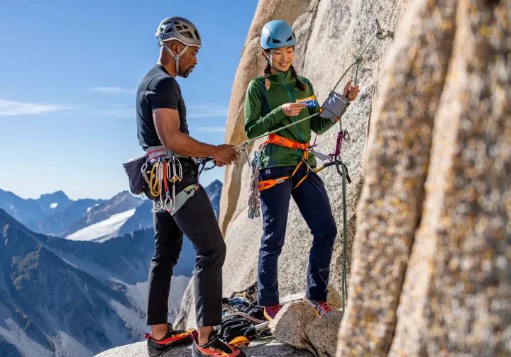 Two diverse alpinists efficiently managing a belay transition on a multi-pitch alpine route, one also stowing micro-trash, embodying efficiency, decision-making, and stewardship which are core to the alpinist's ethos for alpine climbing success.
