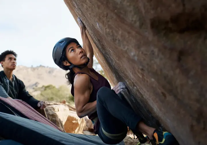 Boulderer paused before a committing move on a highball boulder, looking focused and calm, managing fear with visible crash pads and spotter below, illustrating the psychological dimension of bouldering safety.