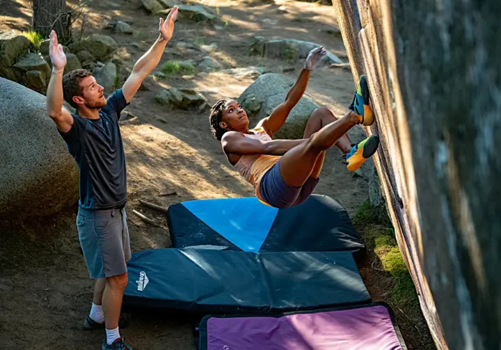 Boulderer taking a controlled fall onto well-placed crash pads with an attentive spotter guiding the landing, preventing bouldering injuries.