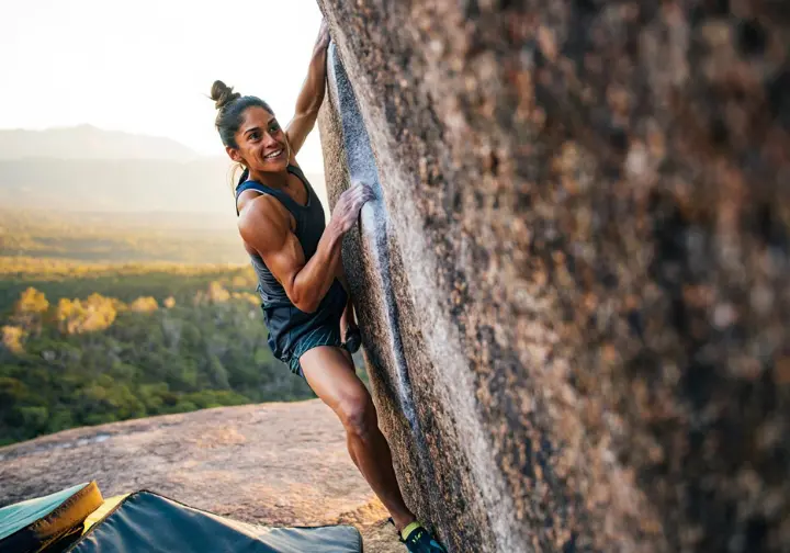 Female boulderer joyfully topping out an outdoor boulder problem at golden hour, showcasing the physical strength, mental focus, and emotional rewards—the many benefits of bouldering.