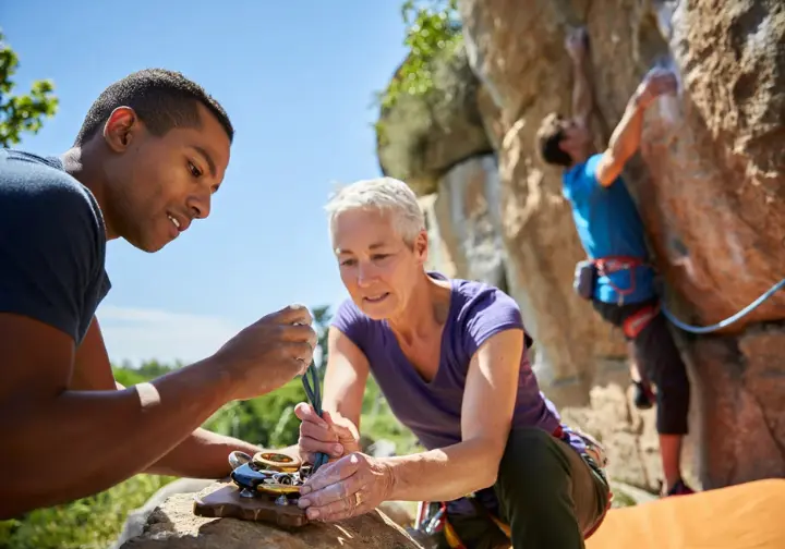 Novice climber practicing trad gear placements at ground level with a mentor, with a scene of them leading an easy trad route in the background, illustrating the learning and progression journey.