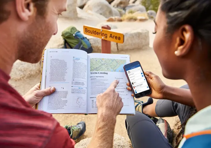 Two diverse climbers using a physical guidebook and a smartphone app to find their way to outdoor bouldering locations, illustrating available resources.