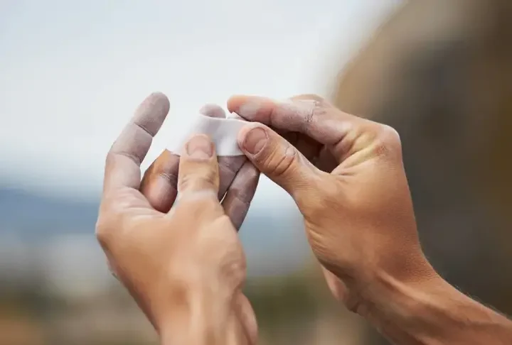 A climber's hands applying white rock climbing finger tape to a finger, illustrating skin protection and pulley support techniques.
