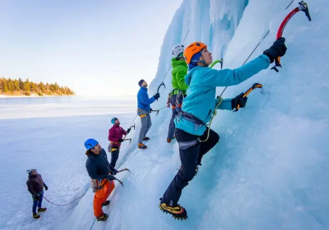 Diverse ice climbers ascend a towering blue and white ice formation above Lake Superior in Michigan's U.P., demonstrating the region's prime ice climbing.