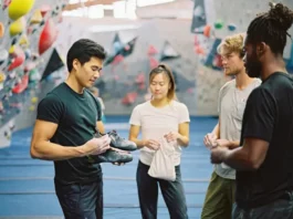 Four diverse climbers in a bouldering gym examining new bouldering gear, including shoes and a chalk bag, representing an essential bouldering gear guide.
