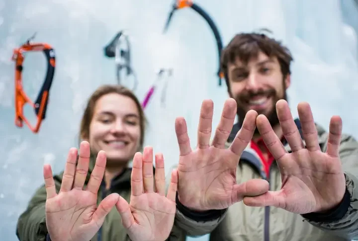 Close-up of two ice climbing gloves on hands, one thin and one thick, against a blurred ice climbing background, demonstrating a multi-glove system for ice climbing.