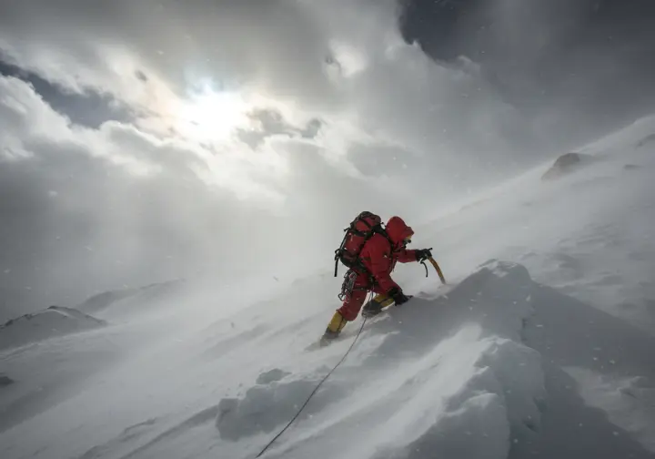A climber in full expedition gear endures the challenge of Aconcagua's difficult and severe weather, with high winds and blowing snow on a steep slope.