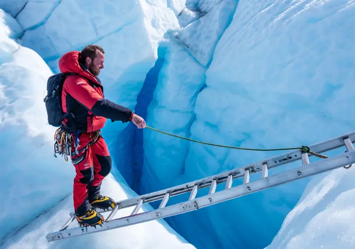 A climber in full gear carefully crosses a crevasse on an aluminum ladder within the treacherous Khumbu Icefall on Mount Everest, surrounded by towering seracs under clear daylight.