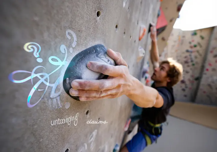A climber's hand on a towering artificial climbing wall, metaphorically untangling the various "world's tallest" claims about the largest climbing walls.