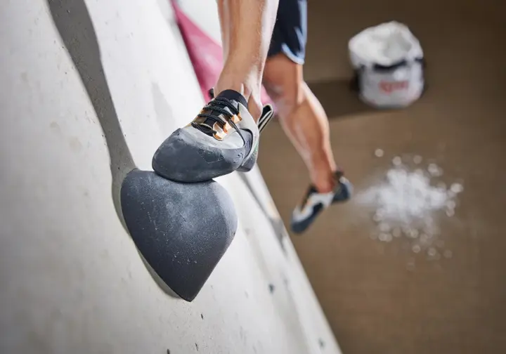 A climber's feet in neutral climbing shoes on an indoor bouldering hold, with a chalk bag nearby, representing the bare minimum bouldering gear for indoor climbing.