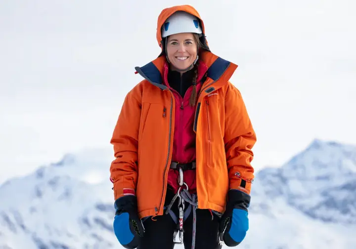 An ice climber dressed in a complete layering system, including a belay parka and multi-glove system, against a snowy mountain backdrop.