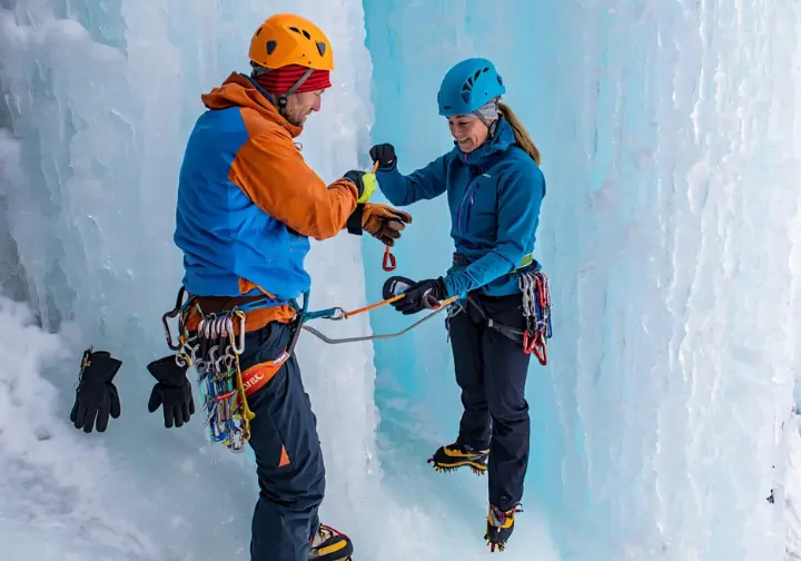 Two ice climbers at a belay station, actively swapping and managing different pairs of gloves on an ice wall, demonstrating system mastery.