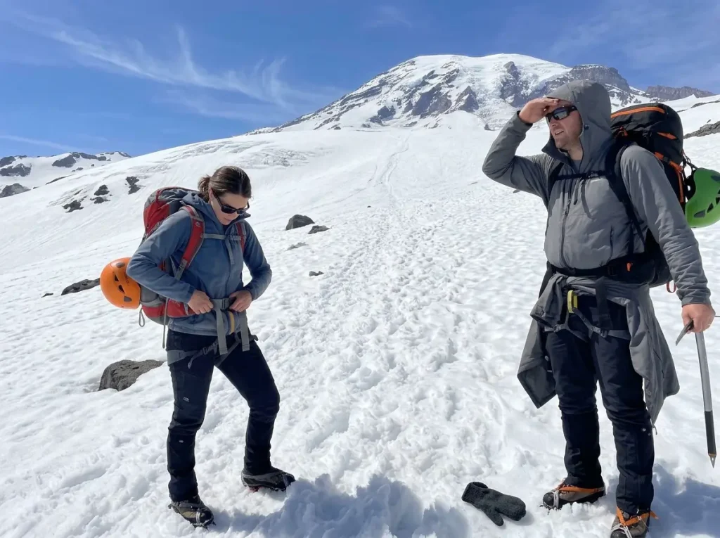 Two climbers pause on bright, firm July snow on Mount Rainier under a hard blue sky, looking up the route.
