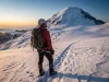 Rainier in July Trades Storms for Crowds and Ladders A mountaineer on a summer snowfield high on Mount Rainier looks up toward the glaciated summit in golden morning light.