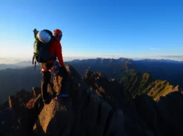 A beginner climber stands on a mountain summit at sunrise, looking out at a vast mountain range, ready for the next challenge.