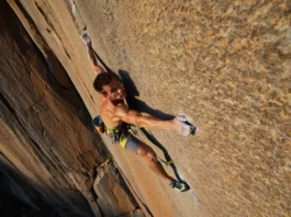 A rock climber demonstrates injury-proof hip and shoulder mobility with a confident high-step and wide reach on a sunny outdoor rock face.