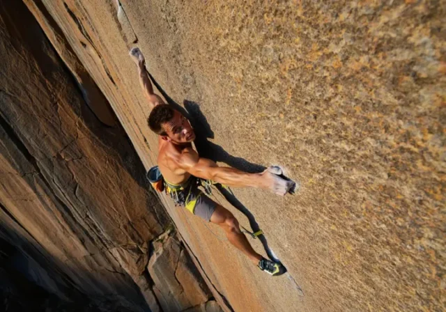 A rock climber demonstrates injury-proof hip and shoulder mobility with a confident high-step and wide reach on a sunny outdoor rock face.
