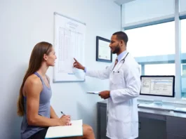 An athletic climber in a doctor's office actively participating in her consultation, holding a notebook while the doctor discusses her care plan.