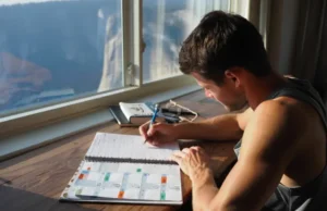 Build Your Climbing Plan: A Periodization Framework A climber sits at a desk creating a periodization training plan in a journal, with a view of a large mountain cliff face through the window behind him.