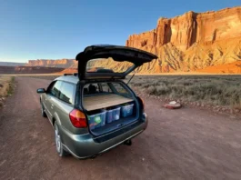 A climber's car is set up for cheap camping in a free, dispersed spot with a view of a massive desert rock climbing cliff at sunrise.