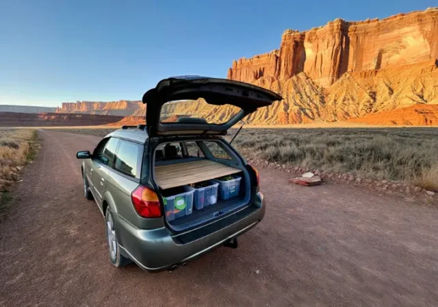 A climber's car is set up for cheap camping in a free, dispersed spot with a view of a massive desert rock climbing cliff at sunrise.