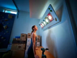 A female climber stands in her modern garage gym, thoughtfully looking up at her new LED training board, representing the choice between Kilter, Moonboard, or Tension.