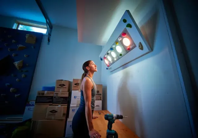 A female climber stands in her modern garage gym, thoughtfully looking up at her new LED training board, representing the choice between Kilter, Moonboard, or Tension.