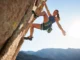 Mobility for Climbers: Unlock High-Steps & Heel-Hooks A female climber demonstrates an advanced mobility move, performing a high-step on a sunny outdoor rock wall, showcasing flexibility and strength.