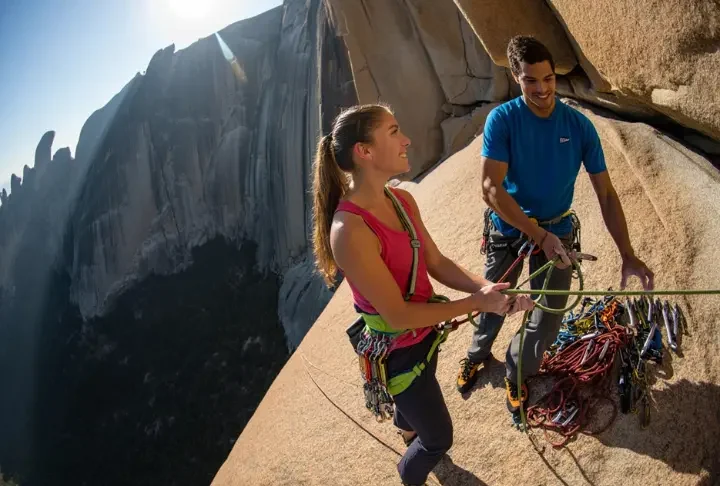 Two competent climbers at a sunny belay station on a vast granite cliff, organizing their gear and ropes as they prepare for the next pitch of their multi-pitch climb.