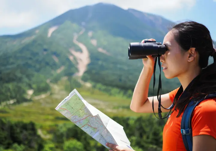 A climber stands using binoculars to analyze the features of a beginner-friendly mountain from across a valley, planning their climb.