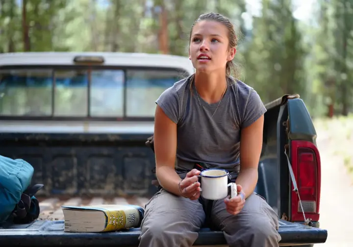 A climber sits thoughtfully on her truck's tailgate, embodying the dirtbag ethos with a coffee mug and a guidebook.
