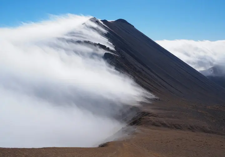 A mountain peak clearly shows how it creates its own weather, with heavy clouds and precipitation on the windward slope and a sunny, dry rain shadow on the leeward slope.